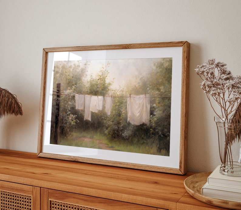 May include: A framed painting of a rural landscape with white clothes hanging on a line to dry. The painting is in a wooden frame and has a soft, muted color palette.