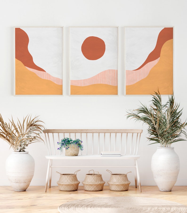 May include: Three framed abstract art prints with a minimalist design featuring orange, red, and white colors. The prints are hung on a white wall above a white bench with a potted plant and three woven baskets.