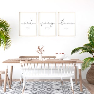 May include: Three framed prints with the words "eat well", "pray often", and "love always" in a minimalist style. The prints are hanging on a white wall above a wooden dining table with white chairs.