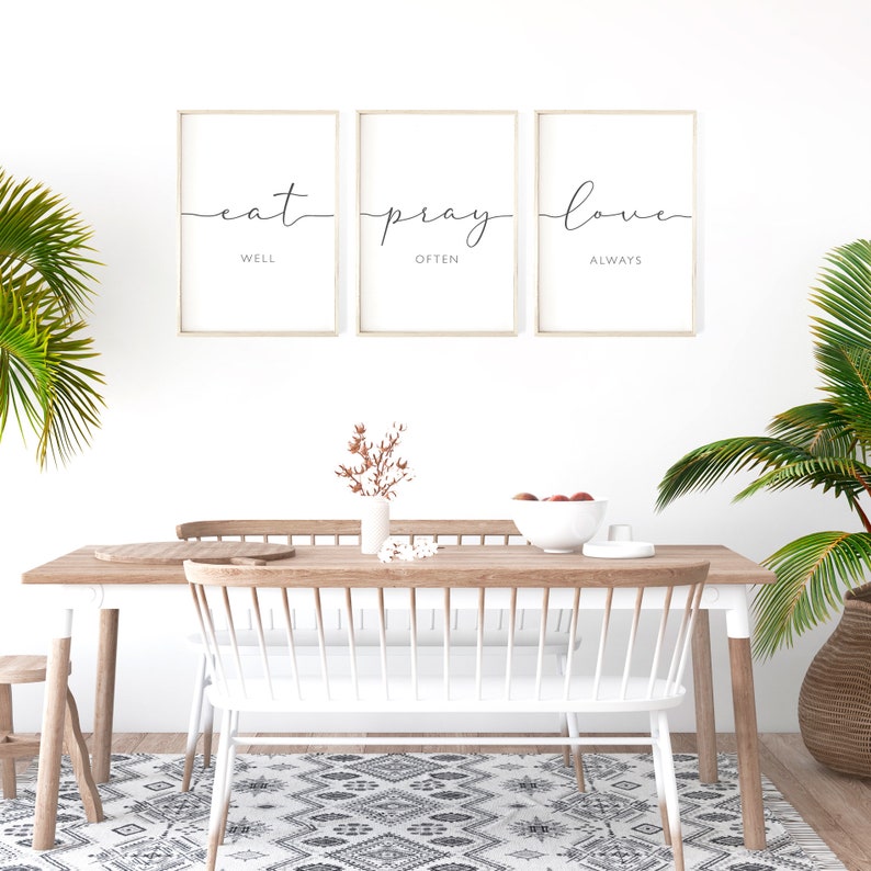 May include: Three framed prints with the words "eat well", "pray often", and "love always" in a minimalist style. The prints are hanging on a white wall above a wooden dining table with white chairs.