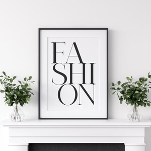 Peut inclure: Impression typographique en noir et blanc avec le mot "FASHION" dans une police de caract&egrave;res moderne. L'impression est encadr&eacute;e dans un cadre noir et expos&eacute;e sur une chemin&eacute;e blanche.