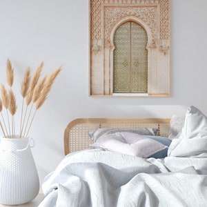 May include: A white wicker headboard with a white and gray patterned pillow on a bed with a white linen duvet. A framed print of a carved archway with a golden door hangs on the wall above the bed. A vase with dried pampas grass sits on the floor next to the bed.