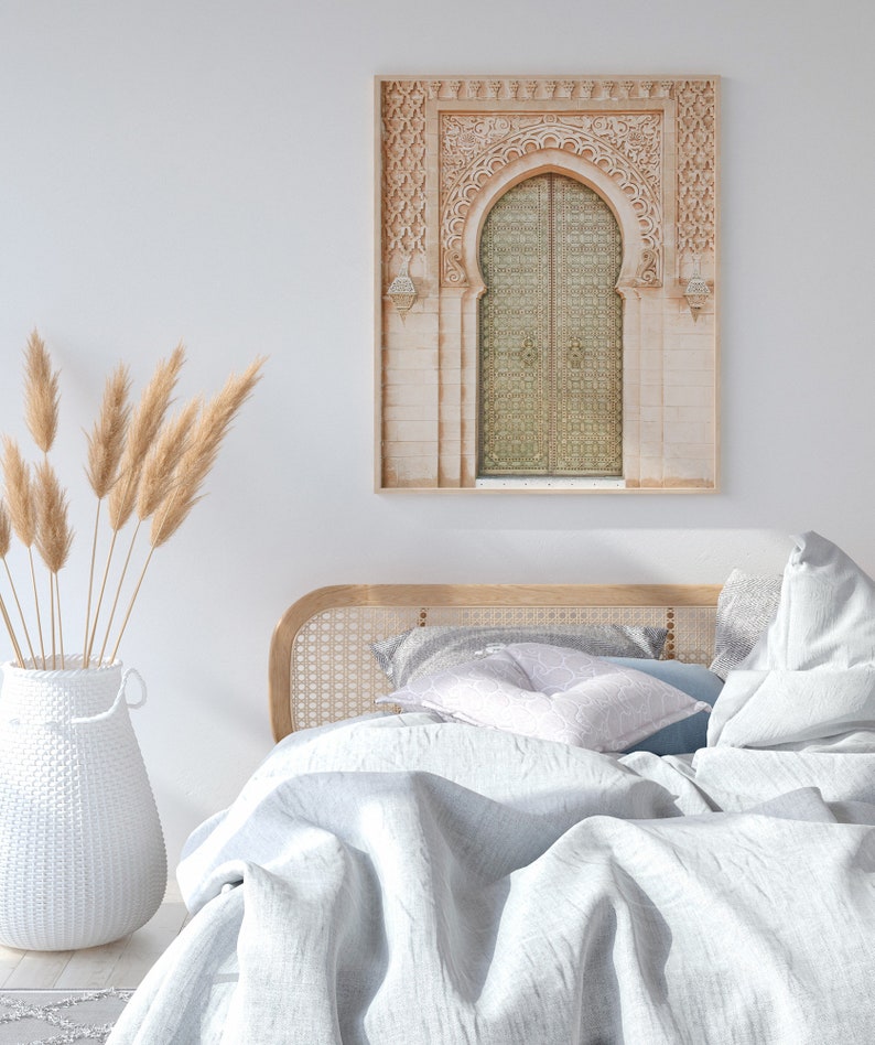 May include: A white wicker headboard with a white and gray patterned pillow on a bed with a white linen duvet. A framed print of a carved archway with a golden door hangs on the wall above the bed. A vase with dried pampas grass sits on the floor next to the bed.