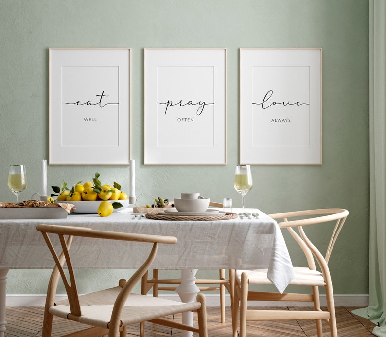 May include: Three framed prints with white backgrounds and black text on a light green wall. The prints read "eat well", "pray often", and "love always". The prints are hanging above a dining table with a white tablecloth and two wooden chairs.