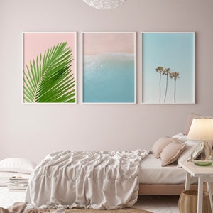 May include: Three framed prints on a pale pink wall above a bed with a white linen duvet. The prints are of a palm leaf, a beach scene with blue water and white sand, and palm trees against a blue sky.