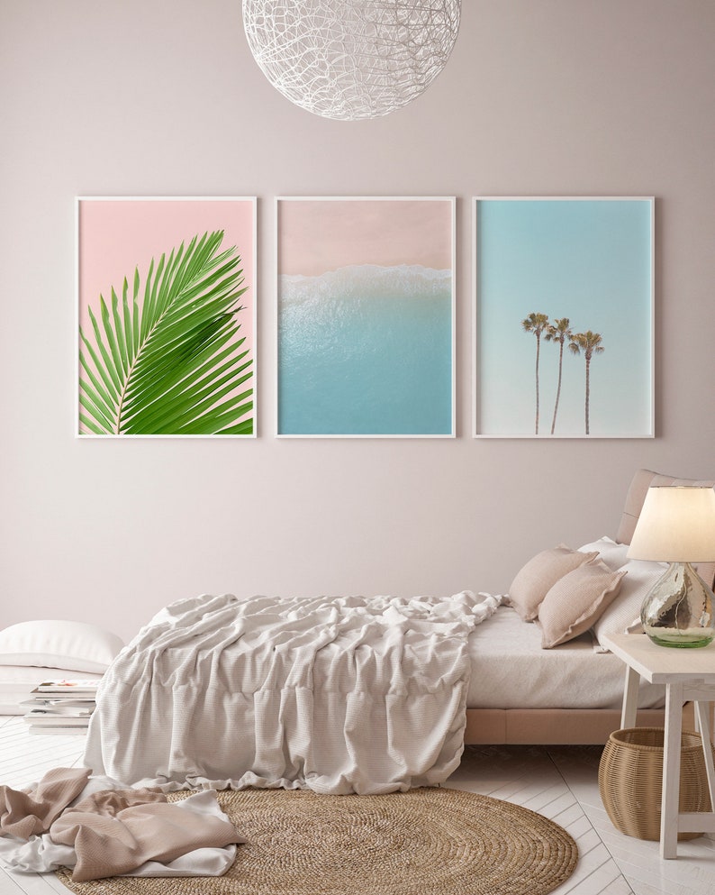 May include: Three framed prints on a pale pink wall above a bed with a white linen duvet. The prints are of a palm leaf, a beach scene with blue water and white sand, and palm trees against a blue sky.