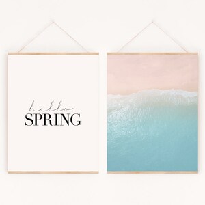 May include: Two framed prints hanging on a white wall. The print on the left is white with black text that says "hello SPRING". The print on the right is a photograph of a beach with blue water and pink sand.