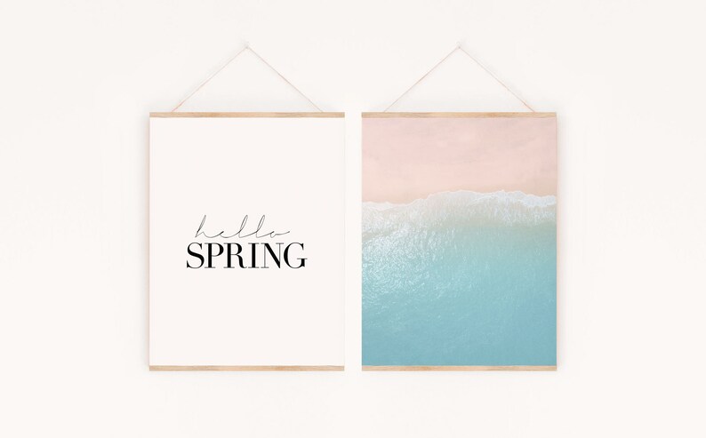 May include: Two framed prints hanging on a white wall. The print on the left is white with black text that says "hello SPRING". The print on the right is a photograph of a beach with blue water and pink sand.