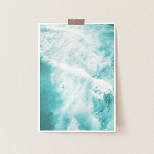 May include: An aerial view of a turquoise ocean with white foamy waves crashing. A single person is seen surfing in the water.