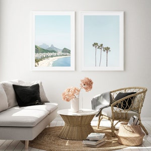May include: Two framed prints of beach scenes. The first print shows a beach with buildings and mountains in the distance. The second print shows three palm trees against a blue sky. The prints are hanging on a white wall above a white couch, a wicker chair, and a wicker coffee table.