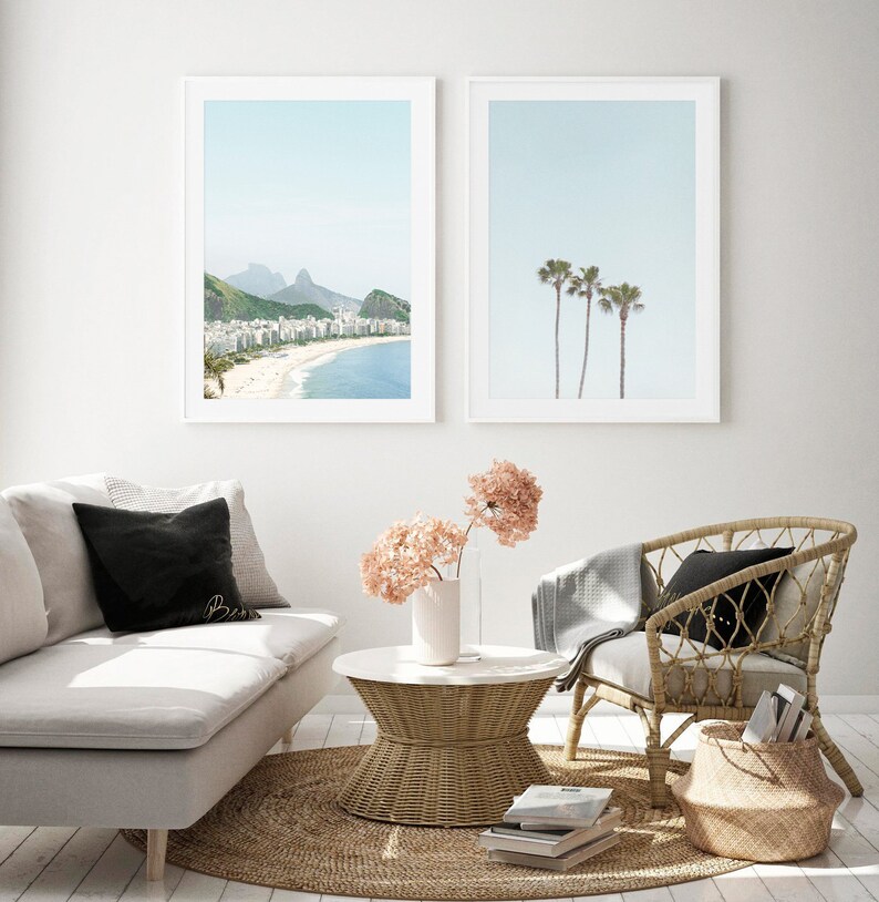 May include: Two framed prints of beach scenes. The first print shows a beach with buildings and mountains in the distance. The second print shows three palm trees against a blue sky. The prints are hanging on a white wall above a white couch, a wicker chair, and a wicker coffee table.