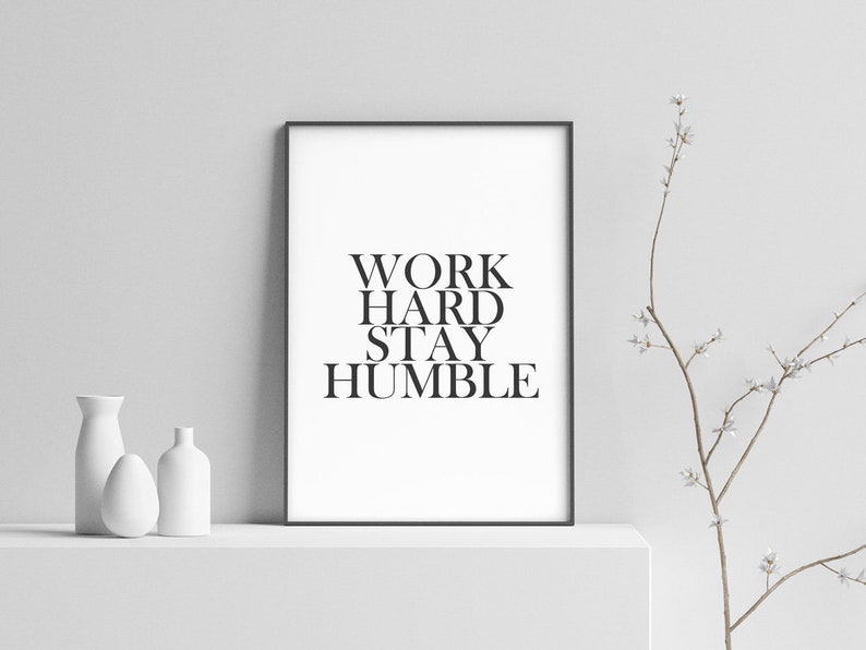 May include: Black and white framed print with the text "WORK HARD STAY HUMBLE" on a white background. The print is sitting on a white shelf with two white vases.