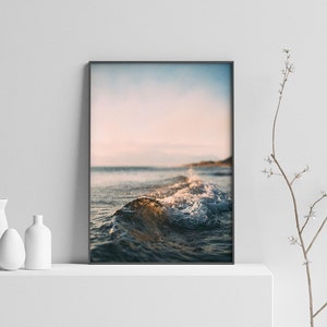 May include: A photograph of a wave breaking on a beach at sunset. The water is a deep blue and the sky is a soft pink and orange.