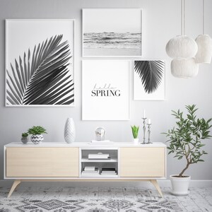 May include: A living room interior with a white media console and three black and white framed prints. The prints feature palm leaves and a beach scene. The text "hello spring" is written on one of the prints. The console is decorated with a vase, a plant, and a decorative object.