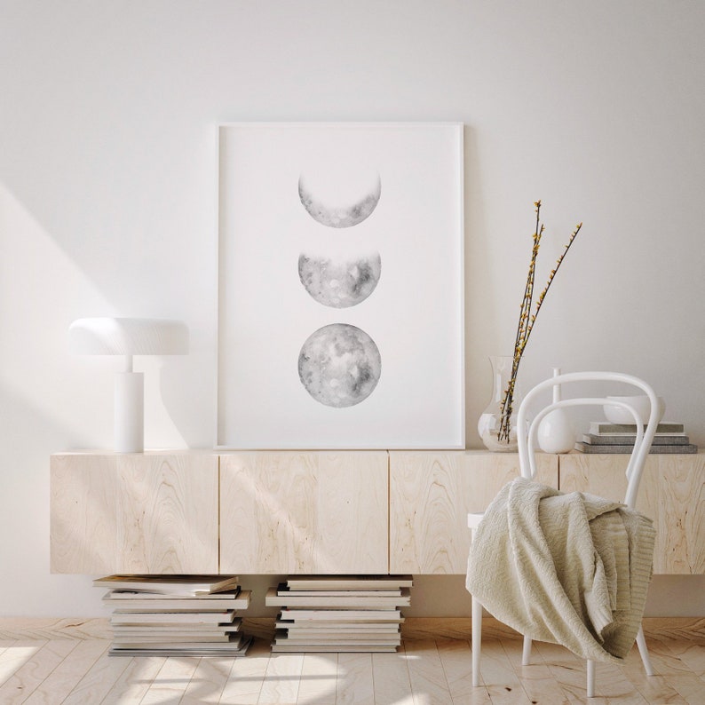 May include: A white framed print with a watercolor illustration of three moon phases in shades of gray. The print is hanging on a white wall above a light wood cabinet with a white lamp and a white chair with a white throw blanket.
