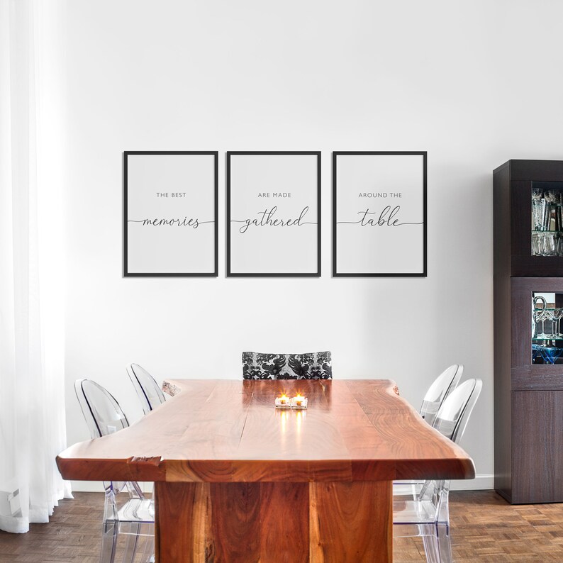 May include: Three framed prints with black borders hang above a wooden dining table. The prints read "THE BEST memories", "ARE MADE gathered", and "AROUND THE table" in elegant script. Clear chairs surround the table.