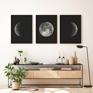 May include: Three black framed prints of the moon in different phases, hanging on a white wall above a wooden console table with black metal legs. The table is decorated with plants, vases, and candles.