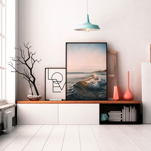 May include: A white wall with a black framed picture of a beach scene with a wave crashing. There is a black framed picture with the word "LOVE" in geometric shapes to the left of the beach scene. A light blue pendant light hangs from the ceiling above the pictures. A wooden shelf with a white cabinet and a black shelf with books and a blue geometric object is below the pictures.