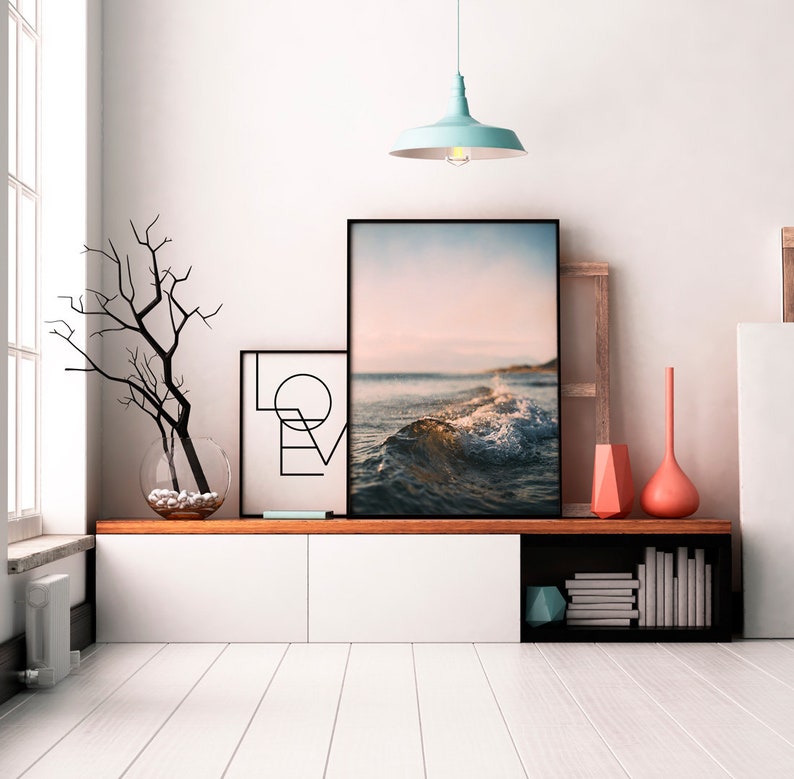 May include: A white wall with a black framed picture of a beach scene with a wave crashing. There is a black framed picture with the word "LOVE" in geometric shapes to the left of the beach scene. A light blue pendant light hangs from the ceiling above the pictures. A wooden shelf with a white cabinet and a black shelf with books and a blue geometric object is below the pictures.