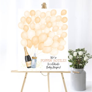 May include: A white sign with a watercolor illustration of champagne bottles and baby bottles with a bunch of peach colored balloons. The sign says "We're Poppin' Bottles to celebrate Baby Harper!"