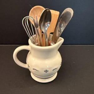 以下が含まれることがあります： 青い花柄のクリーム色の陶器製ピッチャー。中には金属製の泡立て器、小さな金属製のフォーク、木製のスプーン、木製のスパチュラ、金属製のスプーン、金属製のスプレッダーナイフなど、さまざまなキッチン用品が入っています。