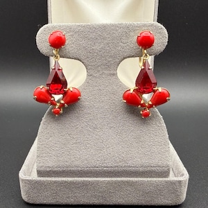 May include: A pair of gold-tone clip-on earrings featuring a round red cabochon top, a central faceted red crystal drop, and three smaller red cabochon accents arranged in a fan-like cluster at the base of each earring piece.