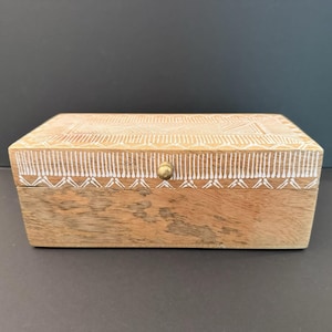 May include: A rectangular wooden box with a hinged lid featuring a small brass knob. The lid is decorated with intricate white painted geometric patterns, including lines and triangles, while the base remains natural wood with visible grain.