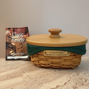 May include: A woven wooden basket with a light brown finish, a matching wooden lid with a central knob, and a dark green fabric liner. A brass-coloured metal plate on the front reads Longaberger 2002 HOSTESS APPRECIATION. Includes a product booklet.