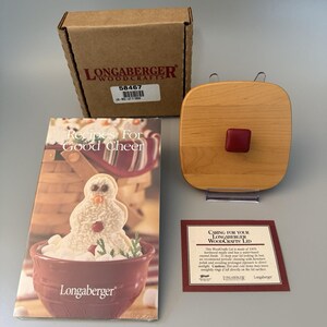 May include: A Longaberger Woodcrafts square wooden lid with a red knob, a cardboard shipping box labeled 58467, a recipe booklet titled Recipes For Good Cheer featuring a snowman, and a printed card with care instructions for the wooden lid.