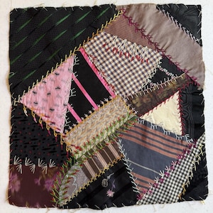 May include: A square crazy quilt block featuring a variety of fabric scraps in black, pink, beige, and grey. The pieces are joined with decorative embroidery stitches in yellow, white, and red, including cross-stitch and feather stitch patterns.