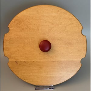 May include: A circular light brown wooden lid featuring a smooth finish and a centered dark red knob. The perimeter of the wooden disc includes four evenly spaced semi-circular cutouts along the edge, providing a distinct notched design.