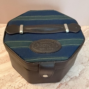 Puede incluir: Un estuche de almacenamiento octogonal con una base de cuero marrón oscuro y una tapa de tela a rayas azules y verdes. La tapa cuenta con un asa de cuero marrón con herrajes plateados y un parche de cuero ovalado grabado con Longaberger Collectors Club.