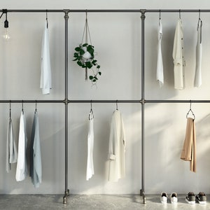 Peut inclure: Un portant à vêtements en métal noir avec deux niveaux de barres suspendues. Le portant est contre un mur blanc et a une plante suspendue au centre. Plusieurs vêtements sont suspendus au portant, dont des chemises, des pantalons et une veste.