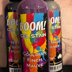 May include: Three bottles of gel stain, labeled "BOOM! Gel Stain". The center bottle is purple and labeled "Gouldian Finch Mauve". The bottle on the left is black and the bottle on the right is silver.