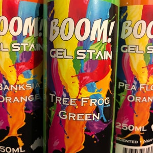 May include: Three cylindrical bottles of gel stain are displayed. The labels feature the word "BOOM!" in white, with "GEL STAIN" below. One bottle is green with "TREE FROG GREEN" text. The other bottles are orange, with "BANKSIA ORANGE" and "PEA FLOWER ORANGE" text.