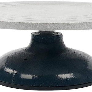 May include: A pottery wheel with a circular, silver-coloured metal top featuring concentric etched rings for centring. The top is supported by a heavy, dark blue, dome-shaped cast iron base that provides stability for rotating clay projects.
