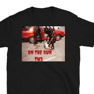 Op de afbeelding: Zwart T-shirt met een grafische afbeelding van een politie-arrestatie bij een rode auto. De tekst "ON THE RUN TW3" is in het rood onder de afbeelding gedrukt. Het shirt is gemaakt van een zacht, comfortabel materiaal.