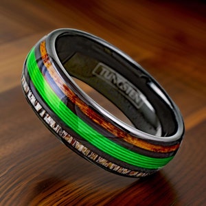 May include: A black tungsten ring with a green and brown wood inlay. The ring is engraved with the word "Tungsten".
