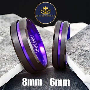 May include: Two tungsten carbide rings with a black finish and a purple inlay. The ring on the left is 8mm wide and the ring on the right is 6mm wide. The rings are on a gray and white textured surface. The text "TUNGSTEN" is engraved on the side of the ring on the left.