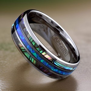May include: A silver tungsten ring with a blue and green abalone inlay. The ring is engraved with the word "TUNGSTEN".
