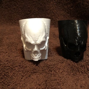 May include: Two 3D printed skull shot glasses, one white and one black. The glasses are sitting on a brown towel.