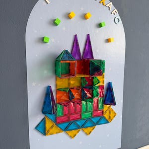May include: A colorful magnetic tile construction set arranged on a light blue background with white stars. The structure includes various geometric shapes in shades of red, green, yellow, orange, and blue. Colorful letters are arranged above the structure.