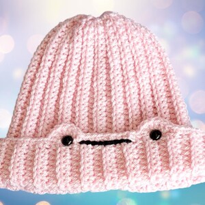 May include: A pink crocheted beanie with black eyes and a black mouth. The beanie has a ribbed texture.