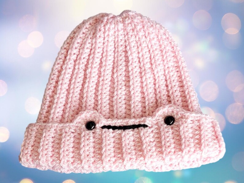May include: A pink crocheted beanie with black eyes and a black mouth. The beanie has a ribbed texture.