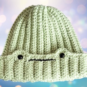 May include: A light green crocheted beanie hat with black button eyes and a black embroidered mouth. The hat has a ribbed texture.