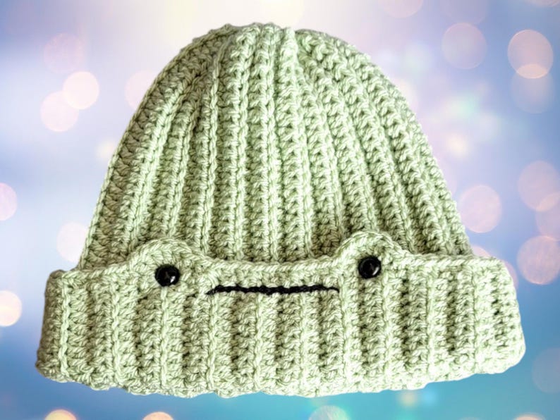 May include: A light green crocheted beanie hat with black button eyes and a black embroidered mouth. The hat has a ribbed texture.
