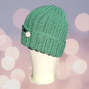 May include: A green crocheted beanie with a pink flower and black button detail. The beanie is on a white mannequin head.