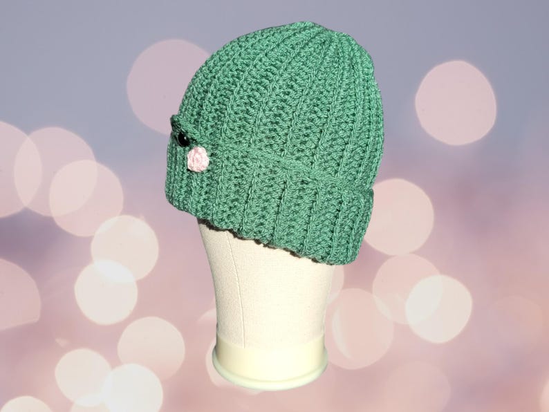 May include: A green crocheted beanie with a pink flower and black button detail. The beanie is on a white mannequin head.