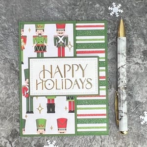 May include: A rectangular greeting card featuring a pattern of toy soldiers in green and red uniforms on a white background, with a central white label reading HAPPY HOLIDAYS in gold text, next to a white and grey marble-patterned ballpoint pen.