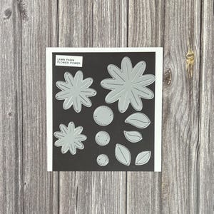 May include: A set of silver metal craft dies on a black backing card labeled Lawn Fawn Flower Power. The collection includes two large flower shapes, three circular centers with stitched details, and four leaf shapes of varying sizes and designs.